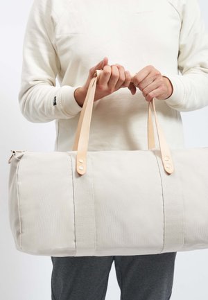 Dufflebag i lys beige lærred med to brune læderstropper, med metal hardware og lynlåslukning, holdt af en person.