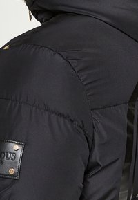 Gros plan sur l'épaule d'une veste matelassée noire avec un écusson de marque en cuir et des boutons-pression dorés.