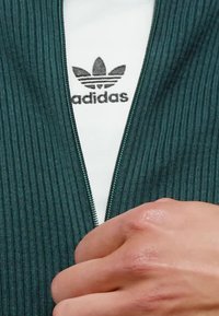 Hand håller i dragkedjan på en grön ribbad jacka, delvis uppdragen för att visa en vit Adidas-tröja med svart logotyp under.