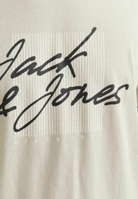 Lichtgrijs T-shirt van katoen, met een groot zwart scriptlogo en een verticale gestreepte achtergrondontwerp. De tekst luidt "SINCE 1990."