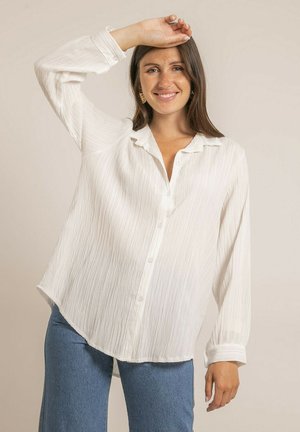 Femme portant un chemisier blanc texturé à manches longues et un jean bleu, souriante, la main droite levée au-dessus de la tête sur un fond uni.