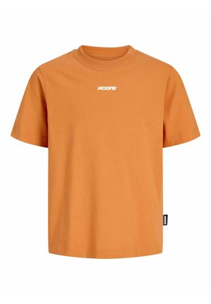 Adolescent portant une casquette noire, un t-shirt orange, un jean gris clair et des bottes beiges, debout et souriant sur fond blanc.