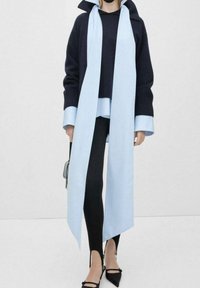 Woman wearing black sweater layered over light blue shirt with matching long scarf, black leggings, and black pointed heels holds small bag.