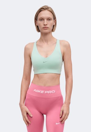 Femme portant un soutien-gorge de sport vert clair et un legging rose Nike Pro taille haute, debout devant un fond uni.