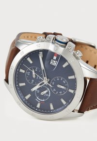 Montre chronographe avec un cadran bleu marine, boîtier en acier inoxydable argenté, bracelet en cuir marron, fenêtre de date et plusieurs sous-cadrans.