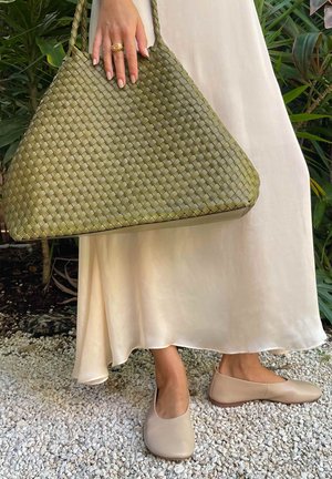 Hand met gouden ring houdt grote groene gevlochten handtas vast; onderbenen dragen beige ballerina's en crèmekleurige satijnen rok op een stenen ondergrond.