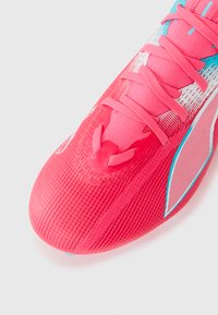 Primer plano de una zapatilla deportiva de color rosa brillante con un upper texturizado, cordones a juego y acentos en azul claro y blanco sobre un fondo blanco.