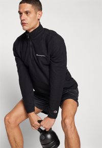 Sudadera negra con cremallera, hecha de un tejido suave, que presenta un pequeño logotipo blanco. Combinada con pantalones cortos negros, el sujeto sostiene una pesa rusa.