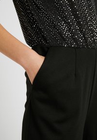 Vêtement noir avec un haut scintillant orné de sequins argentés, associé à un pantalon noir ajusté et lisse avec des poches latérales.