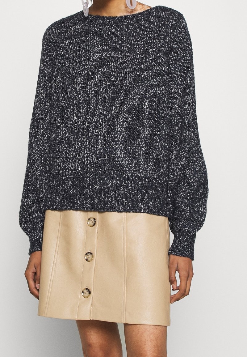 Pull en maille marbrée noir et blanc avec manches ballon, associé à une mini-jupe en cuir beige avec boutons sur le devant.