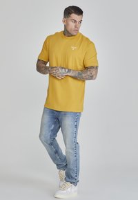Goružastožuta majica kratkih rukava s rebrastim okruglim izrezom, uparena s svijetloplavim trapericama i bijelim tenisicama s teksturiranim vezicama.