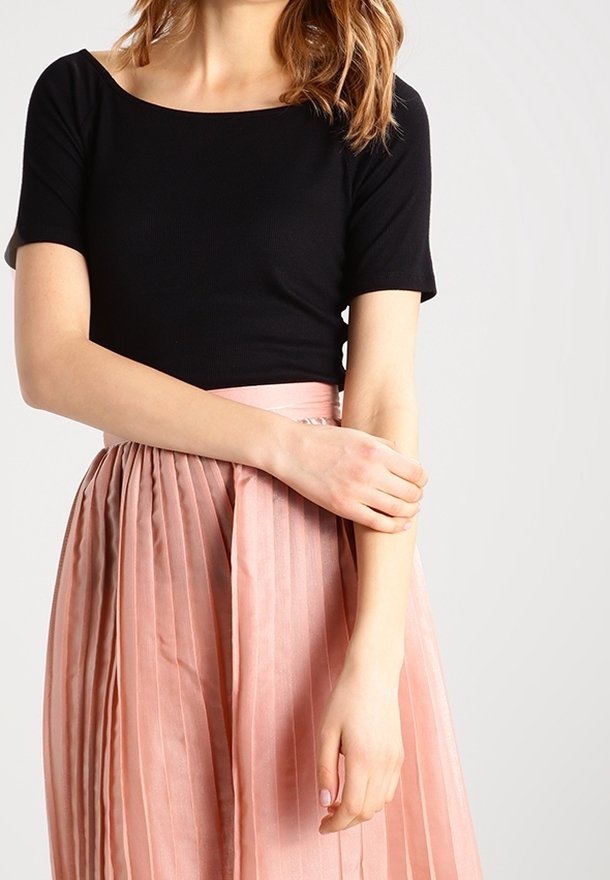 Femme portant un haut noir à manches courtes et une jupe plissée rose taille haute, une main touchant son avant-bras opposé.