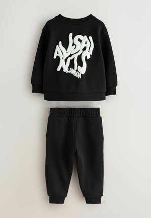 Conjunto de sudadera y pantalones negros con un gran gráfico de texto blanco en la parte frontal. Tela suave, corte holgado, cinturilla el ástica y puños.