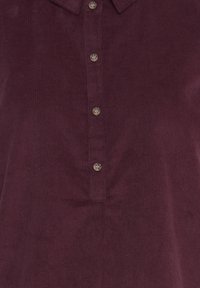Chemise en velours côtelé bordeaux avec quatre boutons visibles et un col pointu, montrée de la poitrine à la taille.