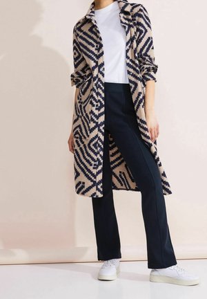 Beige ja laivastonsininen kuvioitu pitkä takki, valkoinen toppi, tummat housut ja valkoiset tenniskenkät. Takissa on geometrinen kuosi ja napillinen etuosa.