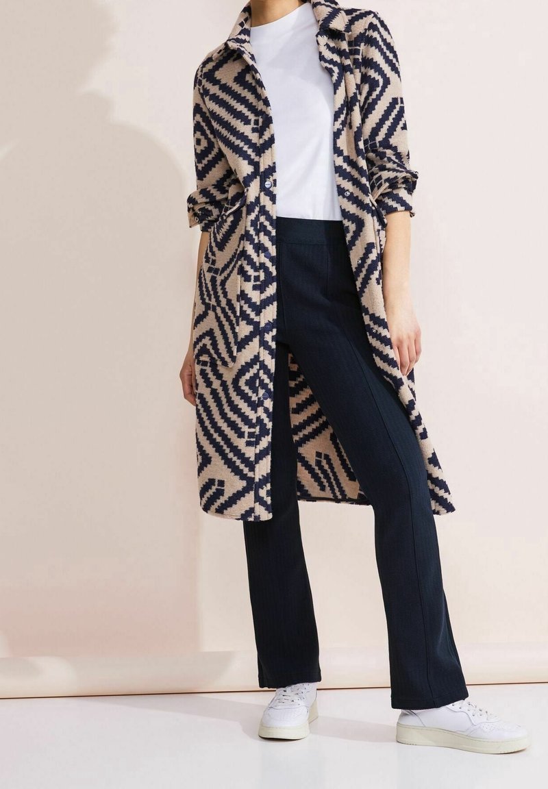 Manteau long à motifs beige et marine, haut blanc, pantalon foncé et baskets blanches. Le manteau présente un design géométrique et une fermeture à boutons.