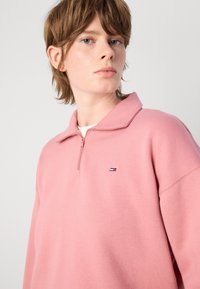 Felpa rosa con zip realizzata in tessuto morbido, caratterizzata da un colletto alto e un piccolo logo sul petto. Vestibilità casual con un design rilassato.