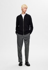 Schwarze Zip-Up-Strickjacke mit Kragen, kombiniert mit grauen karierten Hosen. Das Modell trägt schwarze Slipper. Einfache Hintergrund.