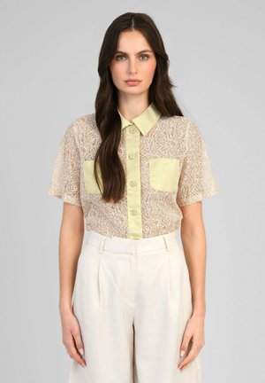 Femme aux longs cheveux bruns portant une chemise en dentelle transparente à manches courtes avec col jaune et poches, associée à un pantalon blanc taille haute.