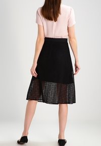 Jupe patineuse en dentelle noire avec un dessus transparent, un motif texturé et une coupe mi-longue. Portée avec un chemisier rose clair.