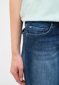 Zils denims bikses ar viegli teksturētu virsmu, ar priekšējo kabatu, jostas cilpām un redzamu metāla pogu aizdari.