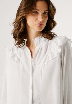 Woman with shoulder-length brown hair wears a white blouse with ruffled shoulders and a high collar, looking slightly to the side.