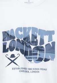 Hvit bomull t-skjorte med stor blå grafisk tekst "HACKETT LONDON," med lagdelt fjellbilde og etableringsdetaljer under.