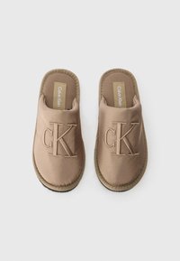 Tan slip-on tofflor med ett mjukt tygöverdrag, präglingen "CK" på ovansidan och en gummisula med en kontrasterande sydd kant.