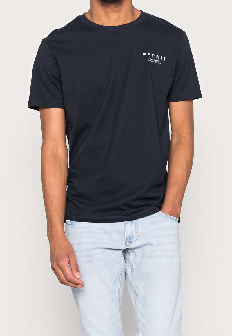 Homme portant un t-shirt bleu marine uni avec un petit logo blanc Esprit sur le côté gauche de la poitrine et un jean bleu clair, debout avec une main dans la poche.