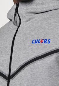 Sudadera gris con cremallera y ribetes negros, y un logo azul. Presenta un cuello alto y una cremallera con cordón. Textura suave, diseño casual.