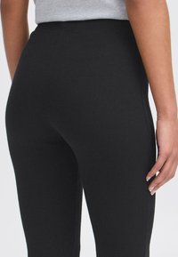 Zwarte geribde legging met een nauwsluitend ontwerp, hoge tailleband en een gladde textuur, gezien vanuit de achterkant met een gedeeltelijk zicht op een hand.