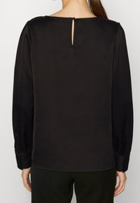 Svart blus med långa ärmar och nyckelhål bak, slät textur och subtil lyster. Ärmsluten har knapprubricering.