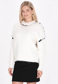 Weißer flauschiger Pullover mit einem gerippten Saum, ausgestattet mit kreisförmigen silbernen Akzenten an den Schultern und dem Halsausschnitt. Kombiniert mit einem schwarzen Rock.