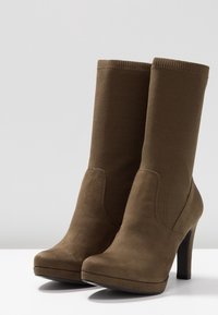 Botas de tobillo de ante marrón con parte superior de punto ajustada, tacón alto y punta redondeada. Presentan una textura suave con un brillo sutil.