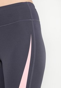 Leggings de alto rendimiento en gris oscuro con una franja diagonal rosa. Textura suave y diseño ajustado con costuras contrastantes.