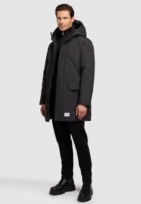 Sort dunjakke med hætte, frontlynlås, to sidelommer og et logo-mærke på den nederste kant. Båret over en sort sweater og bukser.