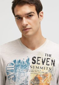 Hellgraues V-Ausschnitt-T-Shirt mit blauem und orangefarbenem Grafikdruck und dem Text "DIE SIEBEN GIPFEL". Weicher, strukturierter Stoff mit lässigem Design.