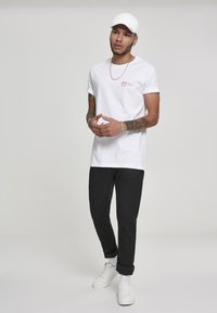 White crew-neck t-shirt with red printed text, black tapered pants, white sneakers, and a white cap. Smooth fabric, relaxed fit.
