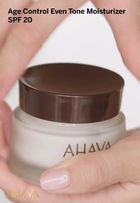 Tarro blanco cremoso con una tapa marrón oscuro, etiquetado como "Hidratante Control de Edad Tono Uniforme SPF 20", sujetado entre los dedos. Acabado brillante en la tapa.