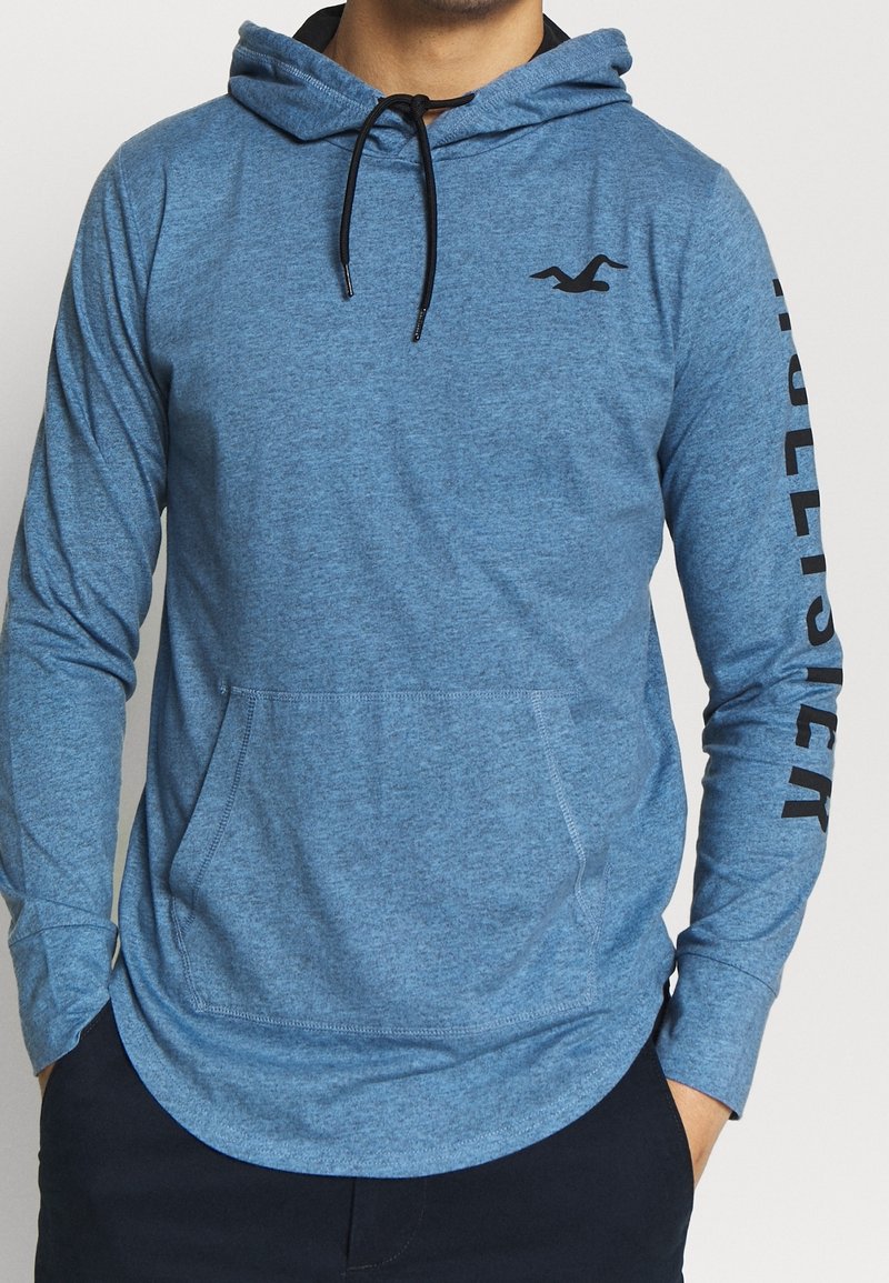 Ljusblå hoodie med lång ärm i texturerad tyg. Har en känguruficka och svart logotyp på bröstet och ärmen.