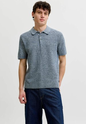 Jeune homme portant un polo gris en maille à manches courtes et un jean bleu foncé, debout devant un fond blanc uni.