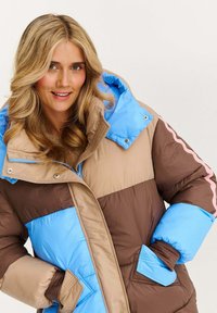Žena s blond vlasmi, oblečená v prešívanej bunde v hnedých, béžových a modrých farbách s kapucňou, usmievajúca sa s rukami v vreckách.