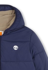 Mørkeblå puffer-jakke med en lys tan fleece-fôr, hette og en hvit trelogo på brystet. Har en quiltet design og myk tekstur.