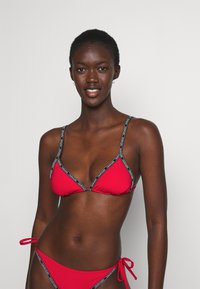 Set bikini rosso composto da un bralette triangolare e slip a vita bassa. Accentuato da cinture nere con il marchio del brand. Tessuto dalla texture liscia.