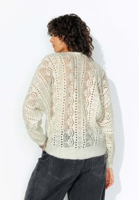 Pull en tricot beige clair avec des motifs ajourés, doté d'un col en V, de manches longues et d'une bordure côtelée. La vue arrière met en avant un design complexe.