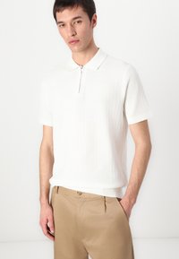 Strikket polo skjorte i off-white med krage, korte ermer og glidelås foran. Bært med lysebrune bukser som fremhever teksturen.