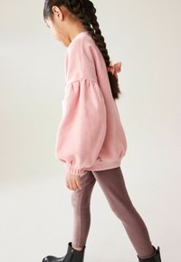 Felpa rosa chiaro con maniche a palloncino, polsini arricciati e dettaglio di un fiocco sulla schiena, abbinata a leggings marroni a coste e stivaletti neri.