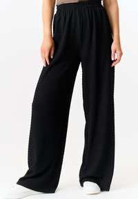Black wide-leg pants made of textured fabric with an elastic waistband. Features a loose fit, reaching the ankles, paired with white sneakers.