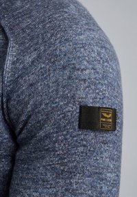Gestrickter Pullover in Blautönen mit strukturiertem Stoff. Besitzt ein schwarzes Etikett an der Schulter mit gelber Schrift und Logo.