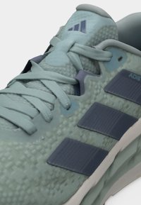 Κοντινό πλάνο ενός αθλητικού παπουτσιού ανοιχτού πράσινου με υφή, με σκούρες πράσινες ρίγες Adidas και κορδόνια στο ίδιο χρώμα, σε λευκό φόντο.